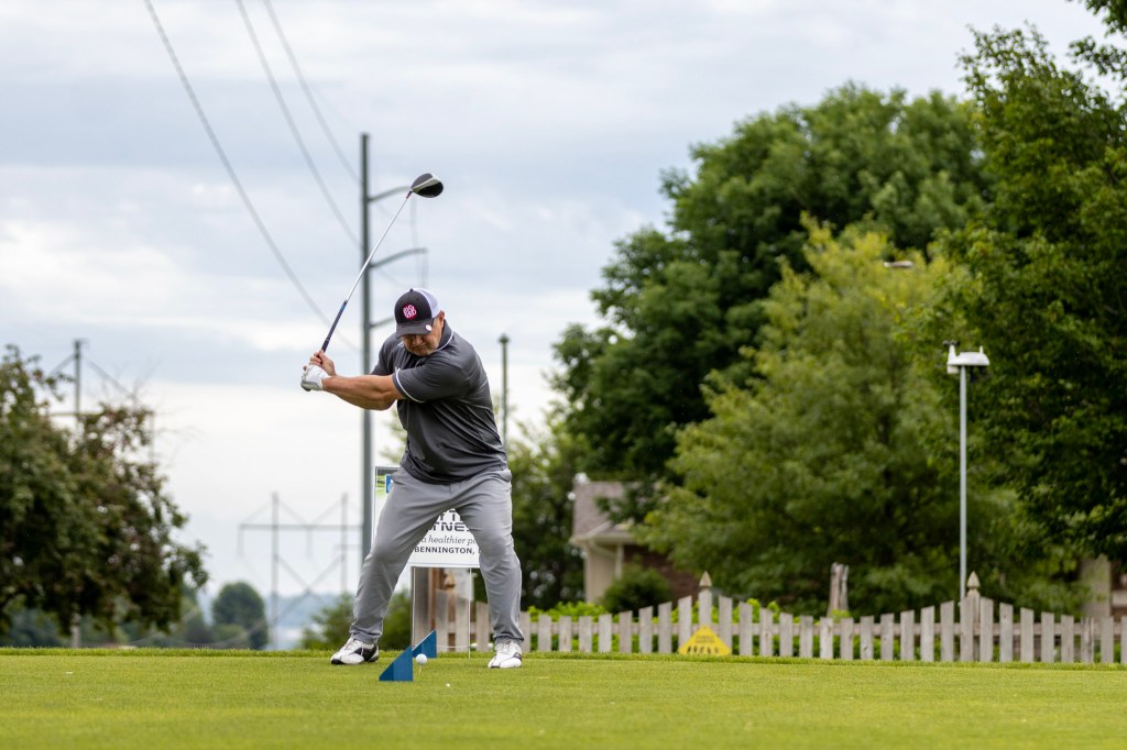 A golfer swings a club on a golf course, poised for a drive. In the background, trees and a fence are visible under a cloudy sky.