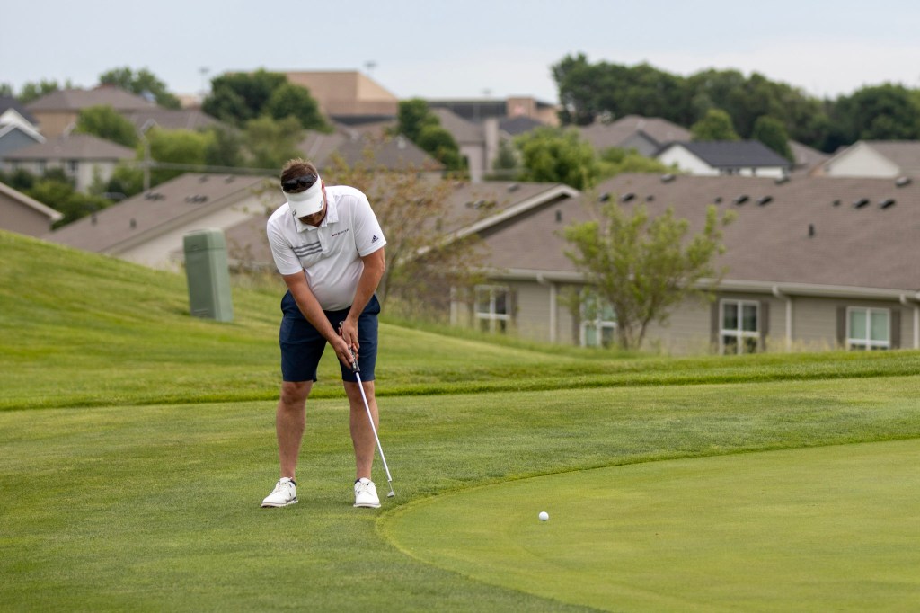 A golfer preparing to putt on a green, with residential buildings and greenery in the background.