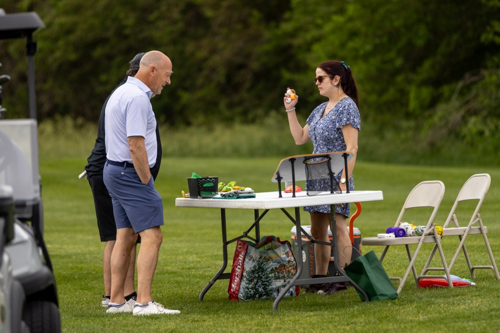Two men conversing while a woman stands at a table, holding an object, with various items on the table in a grassy outdoor setting.