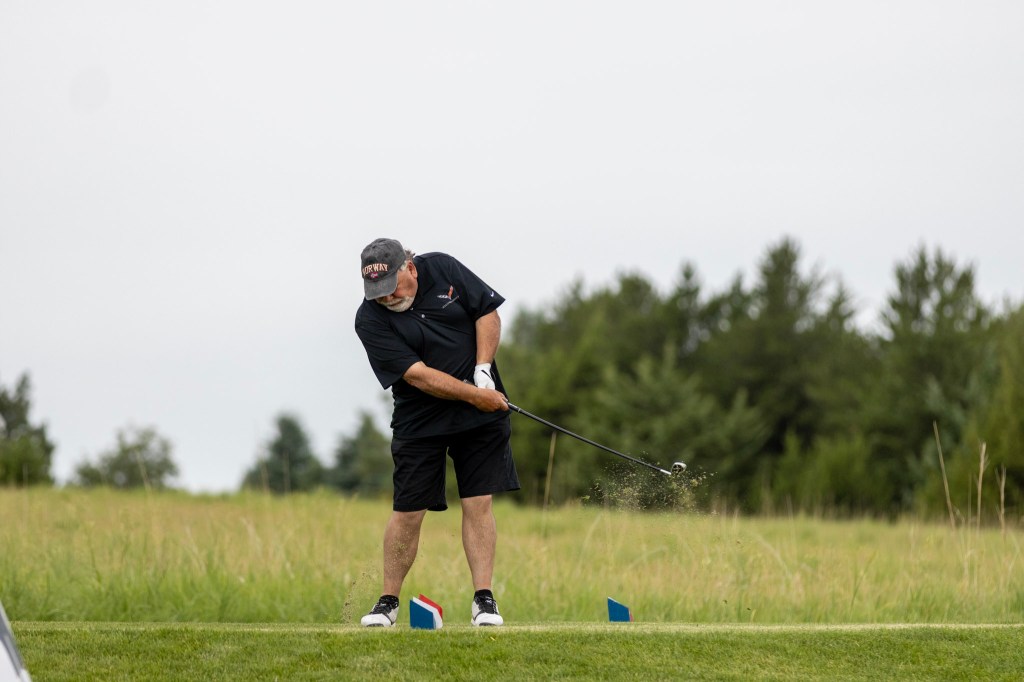 A man in a black shirt and shorts hitting a golf ball on a tee with a lush green background.