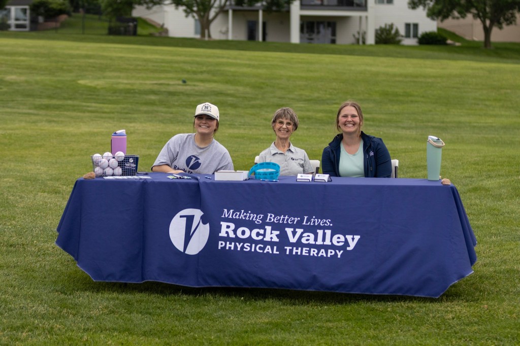 Three women sitting at a table with a blue tablecloth featuring the Rock Valley Physical Therapy logo, surrounded by golf balls and other promotional items, on a grassy field.