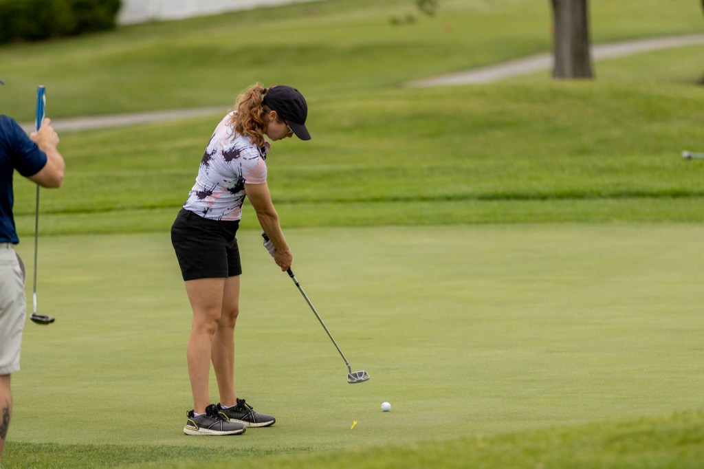 A woman putting on a golf green, wearing a patterned shirt and shorts, with a golf club in hand. A man is partially visible in the background.