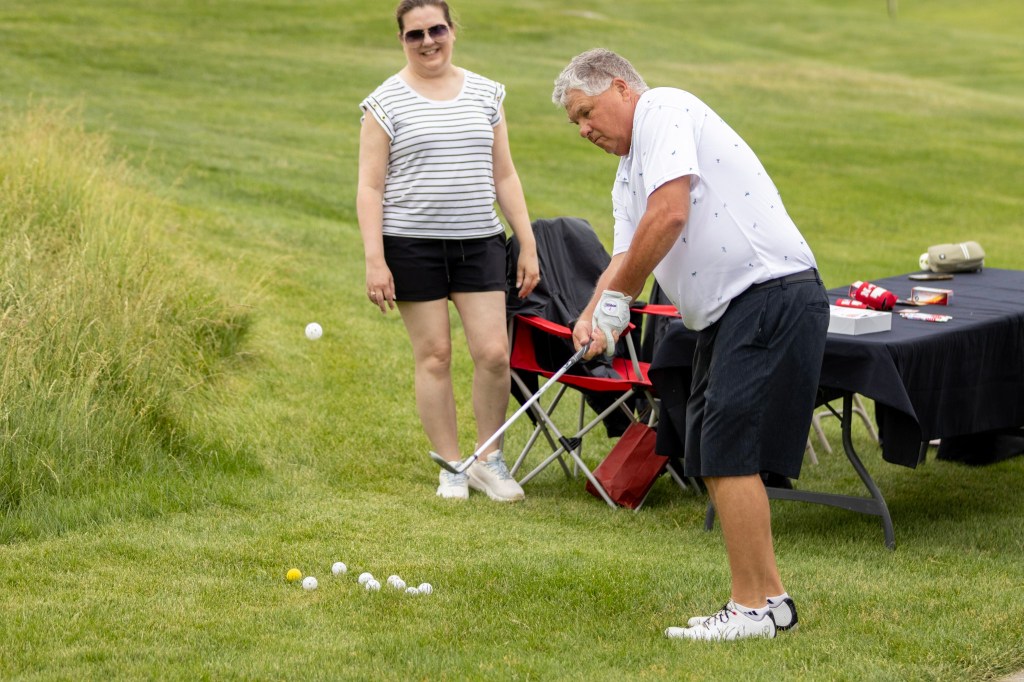 A man in a white polo shirt and black shorts takes a golf swing while a woman in a striped shirt and black shorts watches nearby. Multiple golf balls are scattered on the grass.
