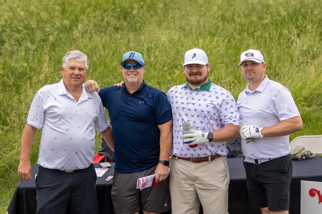 Four men posing together on a golf course, with a grassy background. They are dressed in golf attire, smiling and standing in front of a table with promotional materials.