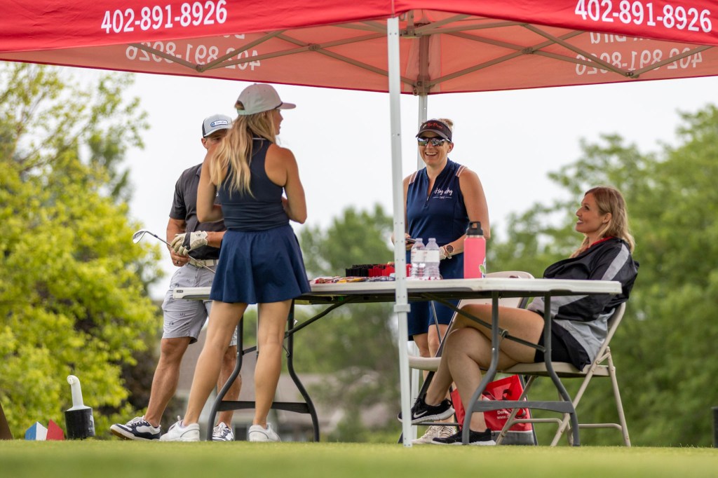 Group of four people interacting under a red tent at a golf course, with one person holding a golf club.