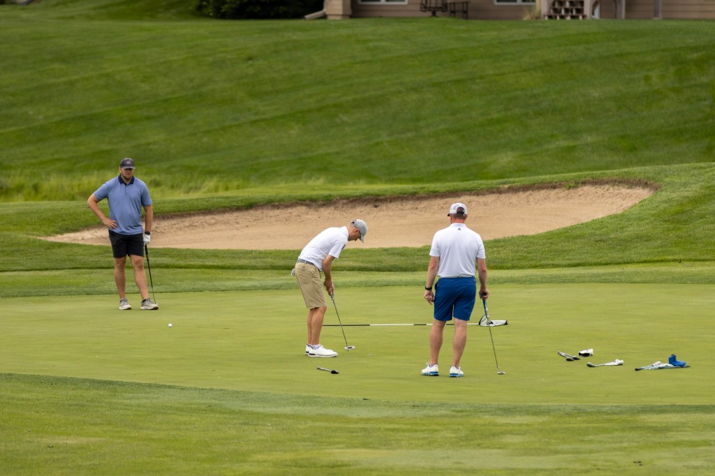 Three men playing golf on a putting green, with a sand trap visible in the background.