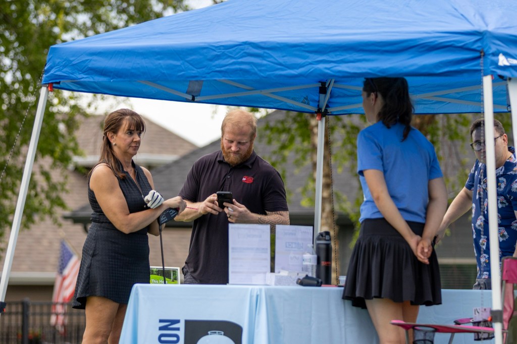 A group of people interacting under a blue canopy at an outdoor event, with a woman in a black dress and a man with a beard looking at a phone, while another woman in a blue shirt stands nearby and two others observe.