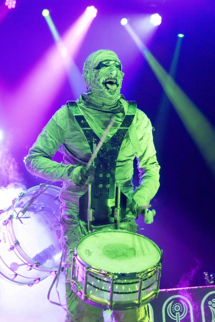 A performer dressed as a mummy plays a snare drum on stage, surrounded by colorful lights and fog.