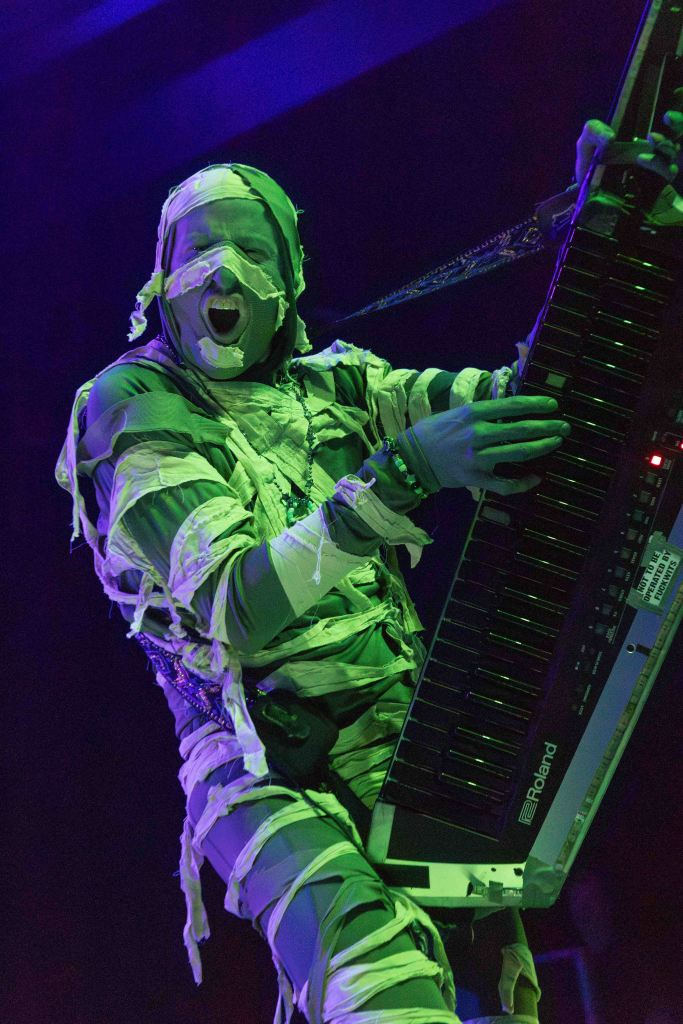 A performer dressed in green and white bandages, playing a keyboard instrument and making an expressive face under purple lighting.