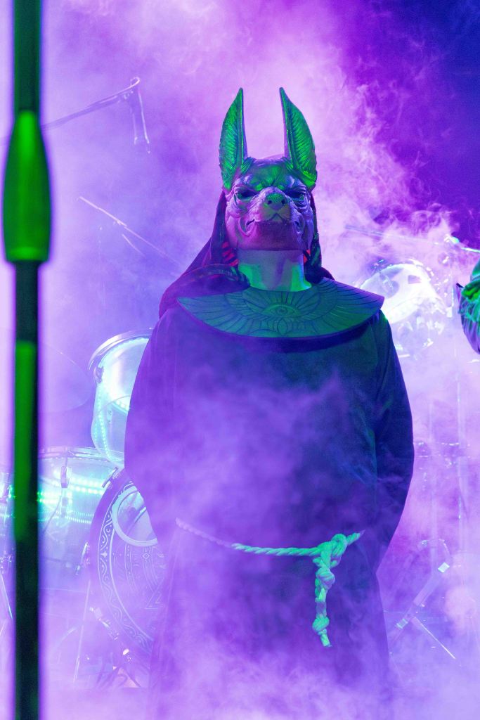 A performer in a dark robe and an elaborate Anubis mask stands amidst colorful smoke on stage.