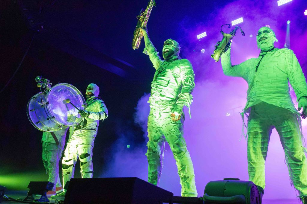 Three performers dressed in green, mummy-like costumes playing music on stage with colorful lights and fog in the background.