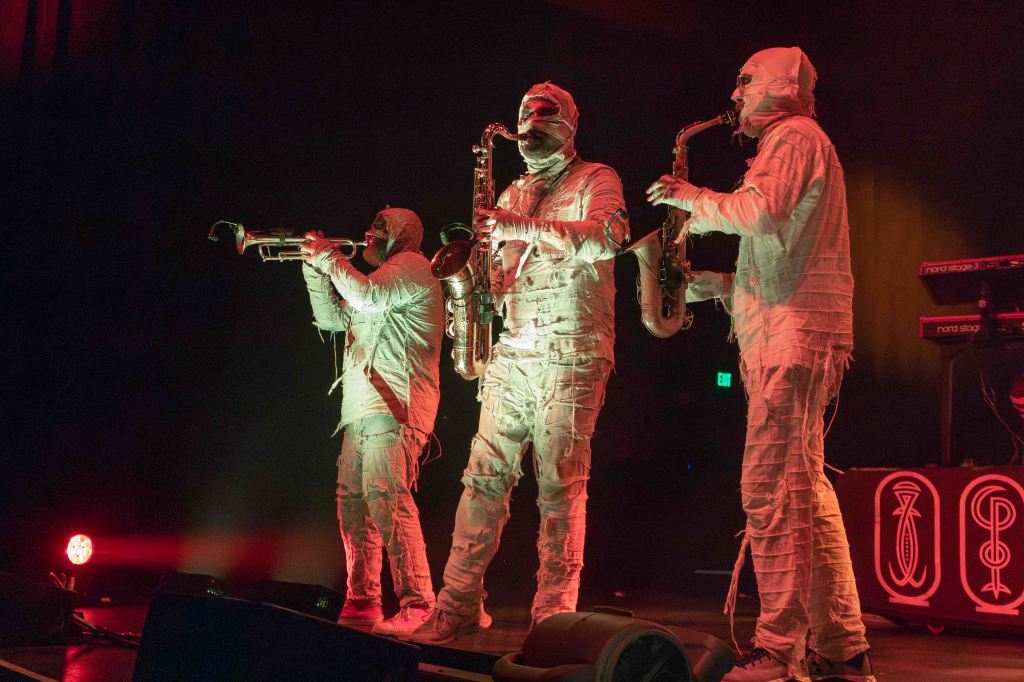 Three musicians dressed in white bandages perform on stage, playing brass instruments. The scene is illuminated with red lighting.