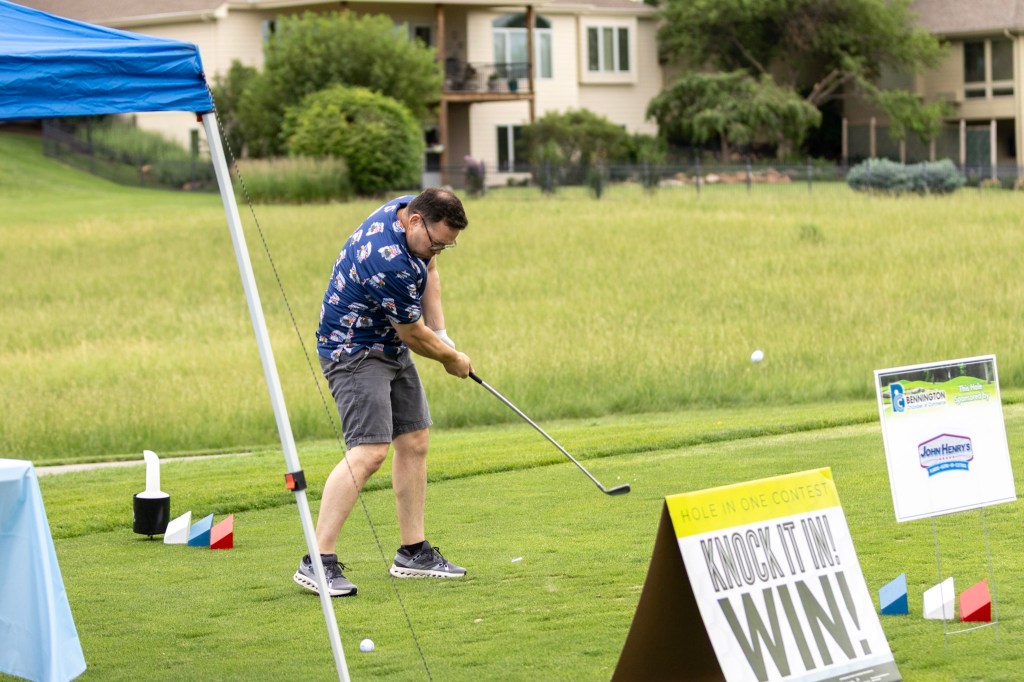 A man in a colorful shirt swings a golf club at a driving range, with a sign promoting a 'Hole in One Contest' nearby.