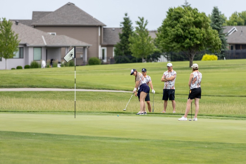 A group of four golfers on a putting green, with one player preparing to putt while the others observe. Residential houses and trees are visible in the background.