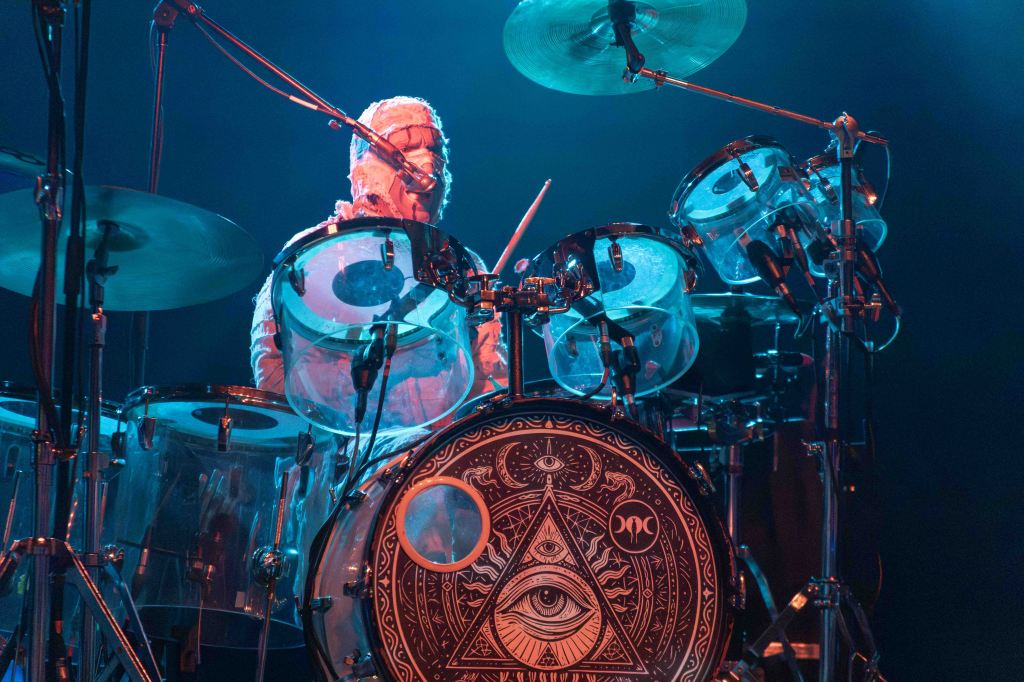 A drummer performing on stage with a drum kit featuring a decorative bass drum with an eye design, surrounded by blue lighting.