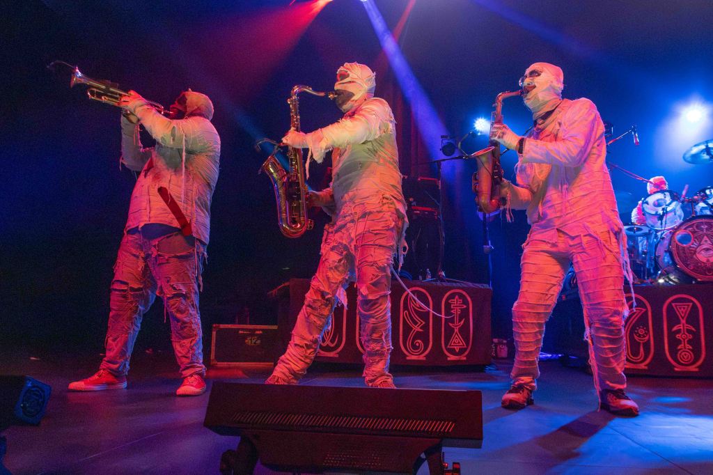 Three musicians dressed in bandages perform on stage with brass instruments under colorful stage lights.
