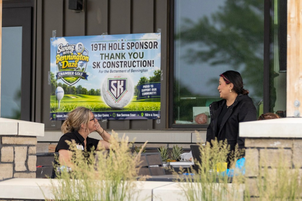 Two women conversing at an outdoor gathering with a banner thanking SK Construction for sponsoring a golf event titled 'Swing into Bennington Daze'.