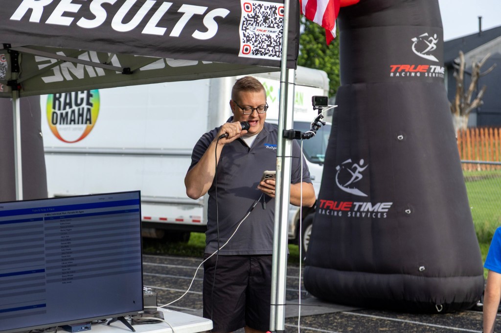 A man speaking into a microphone at a race event, displaying results on a screen, with a True Time Racing Services banner and an American flag in the background.