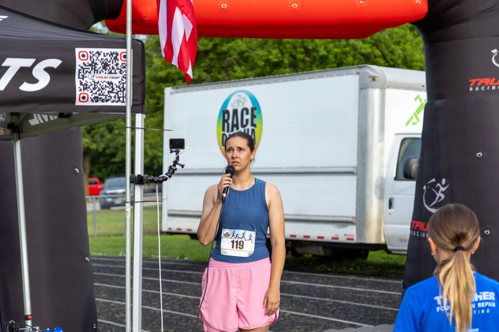 A female speaker stands at a microphone wearing a sports outfit, preparing to address a gathering at a running event. An inflatable arch and an American flag are visible in the background.