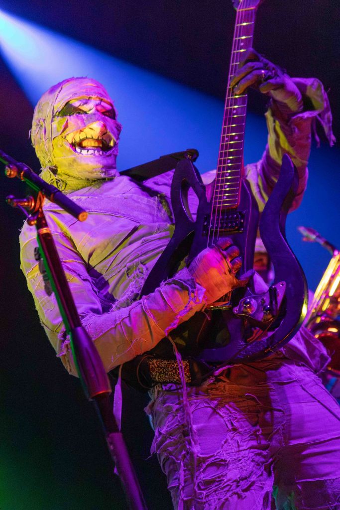 A musician dressed in a mummy costume passionately playing an electric guitar on stage, with colorful stage lighting in the background.