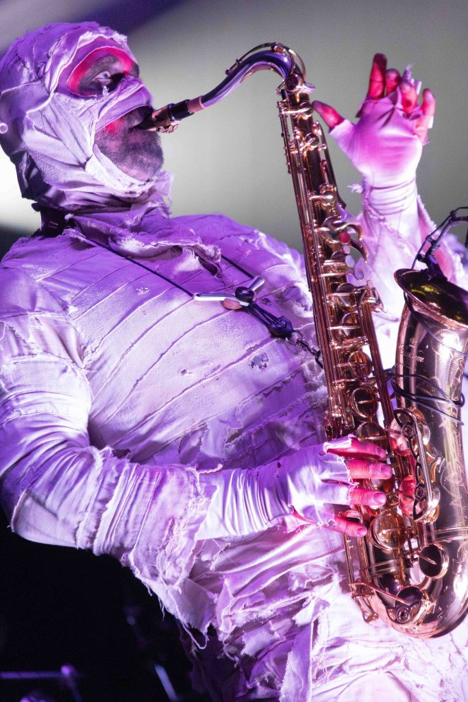 A musician dressed in a mummy costume plays a saxophone on stage, illuminated by colorful lights.