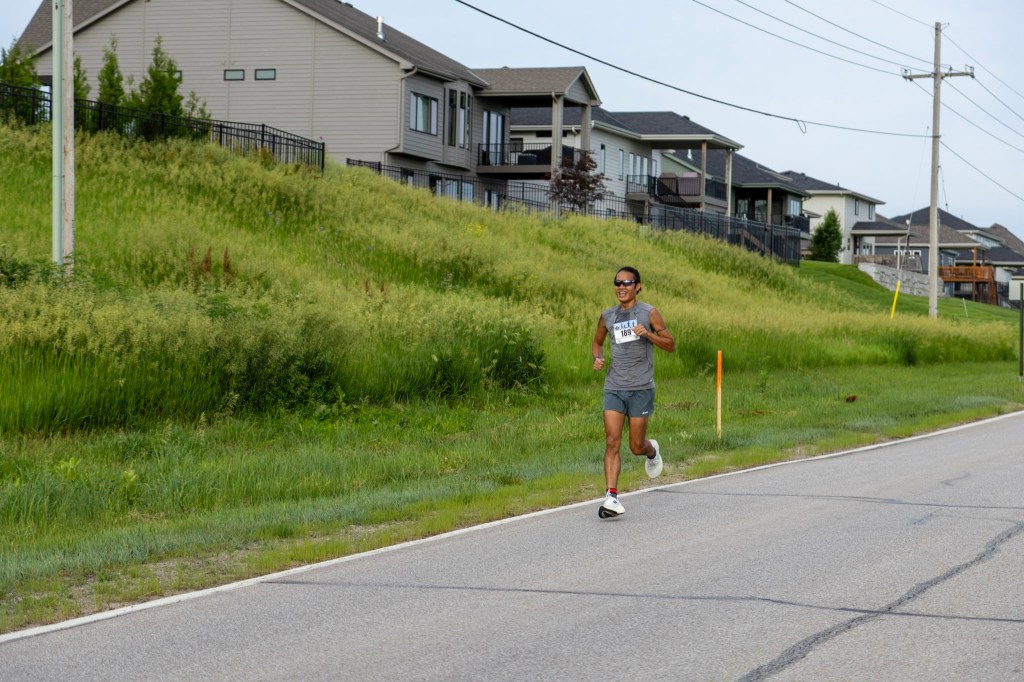 A runner in a gray athletic outfit and sunglasses is jogging along a road with houses and lush green grass in the background.