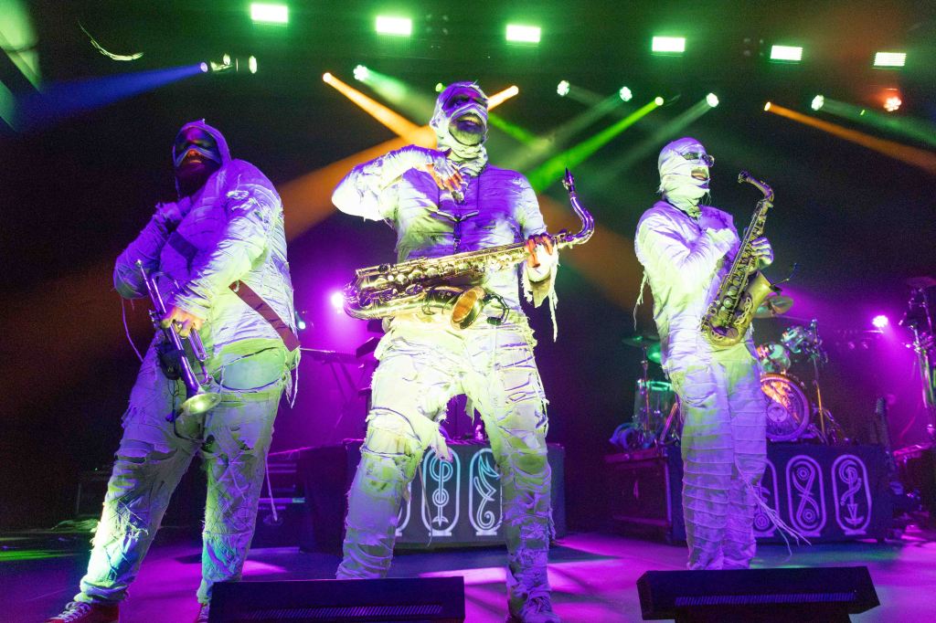 Three musicians dressed in white mummy costumes perform on stage under colorful lights, one playing a saxophone, another holding a trumpet, and the third in a dynamic pose.