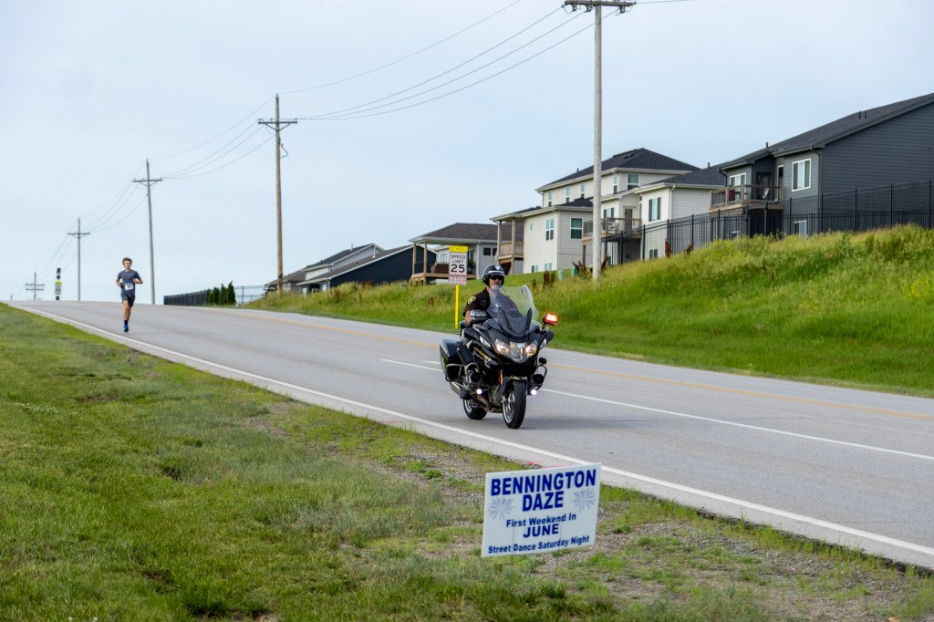 A police officer on a motorcycle assists a runner on a road near residential houses, with a sign for Bennington Daze in the foreground.