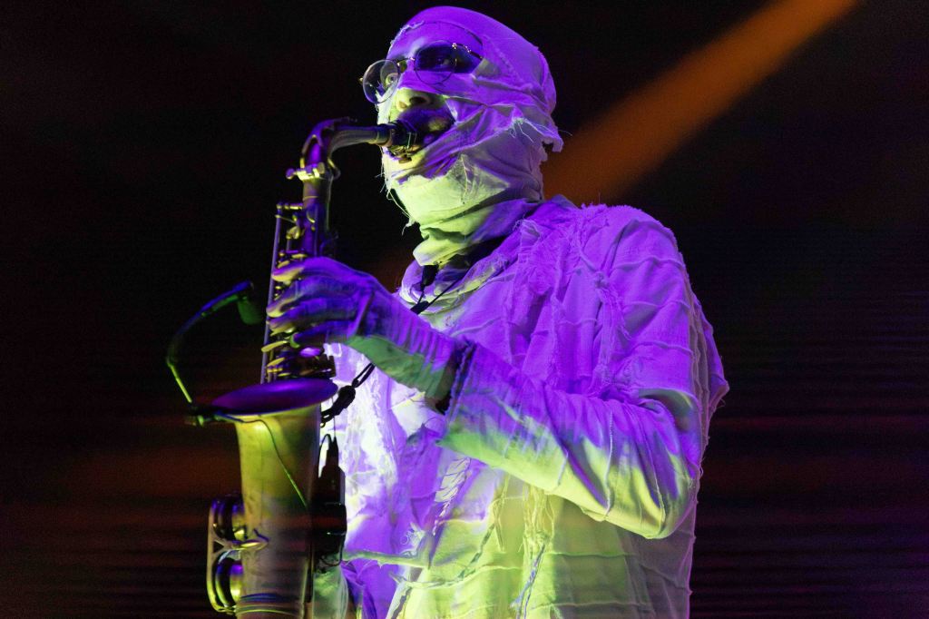 A musician dressed in white and wrapped in cloth plays a saxophone under colorful stage lights.