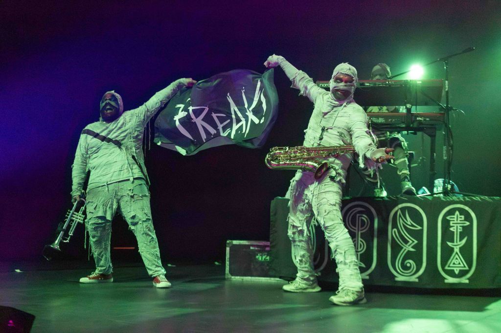 Two musicians dressed in white bandages perform on stage, one holding a trumpet and the other a saxophone, while one brandishes a flag that reads 'FREAK!'