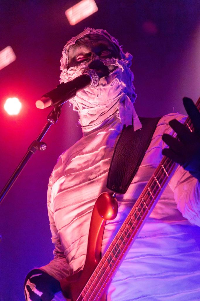 A performer dressed in a mummy costume playing a bass guitar while singing into a microphone, with colorful stage lighting in the background.