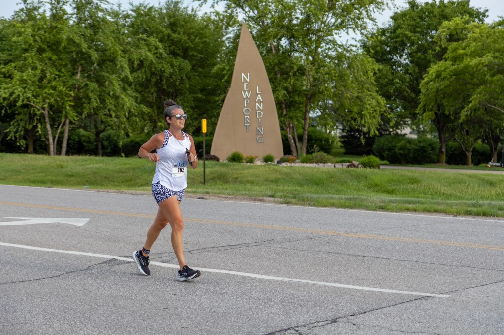 A woman jogging on a road near a sign that reads 'Newport Landing.' She is wearing a tank top and shorts, with sunglasses on, and has her hair tied back.