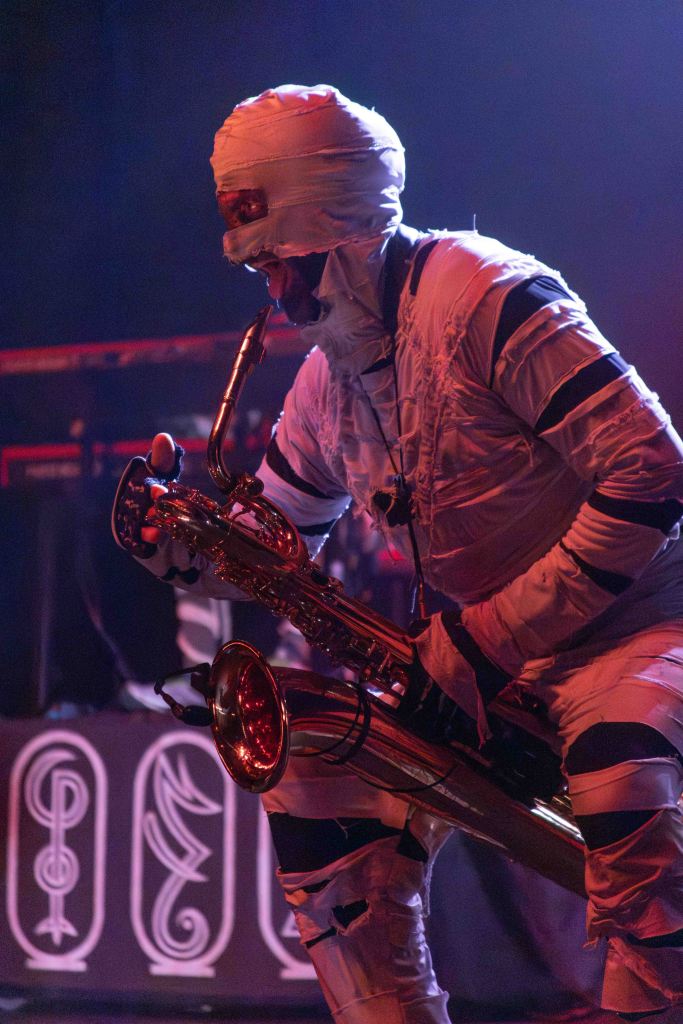 A musician dressed as a mummy playing a saxophone on stage, with dramatic lighting and a patterned backdrop.
