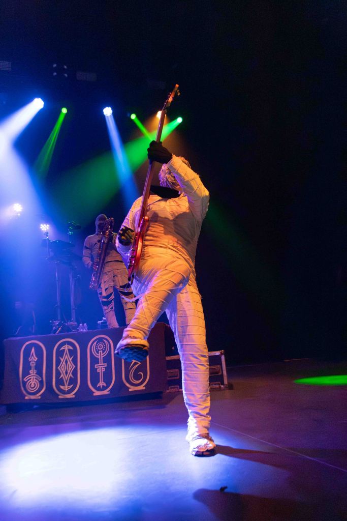 A musician dressed in a white mummy costume playing an electric guitar on stage, with colorful stage lights in the background.