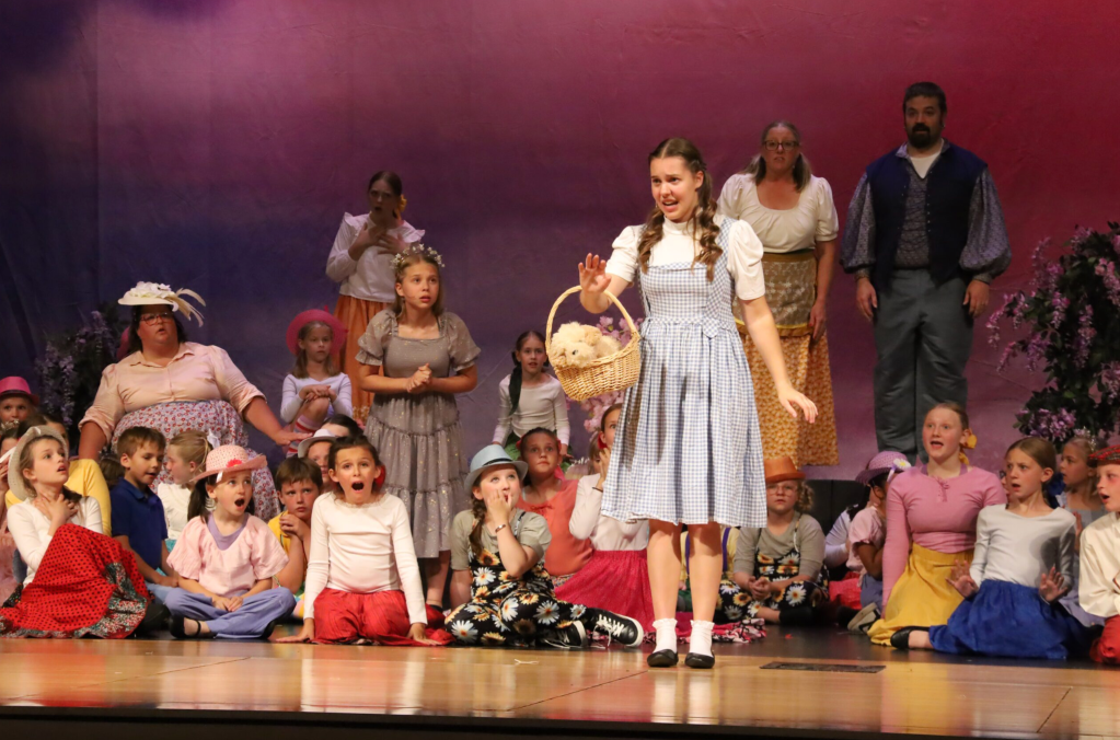 A stage performance featuring a young girl in a blue dress with a basket, surrounded by a group of children and adults in various costumes, all reacting with surprise and excitement.
