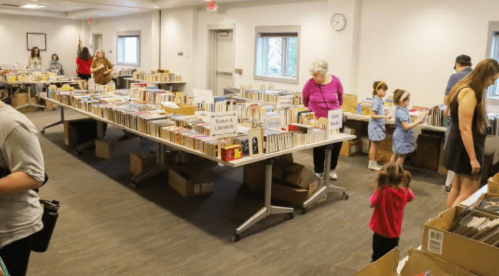 A busy indoor book sale event with several tables filled with books. People of various ages are browsing through the books, including children. Some boxes are placed on the floor for additional storage.