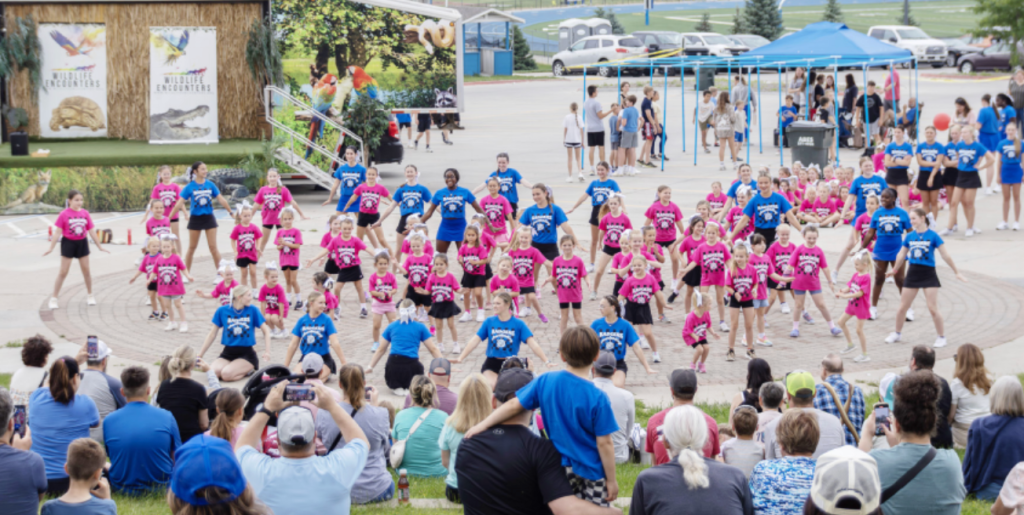 A large group of children in colorful shirts performing a dance routine on a stage, with an audience seated in front of them. The performers are wearing blue and pink shirts, and there are banners in the background featuring wildlife imagery.