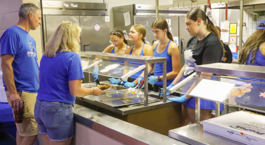 A group of people serving food in a cafeteria setting, with two adults and four teenagers wearing blue shirts. The adults are standing in front of a serving counter, while the teenagers assist behind it, wearing gloves and preparing plates.