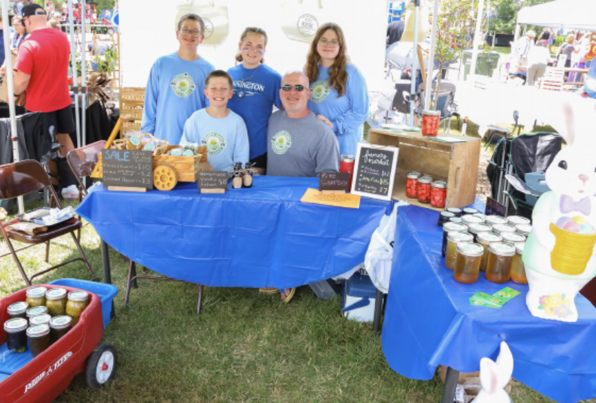 A family of five standing behind a booth at an outdoor market, displaying homemade goods on a blue tablecloth. Various jars of preserves and baked items are visible, along with price signage.