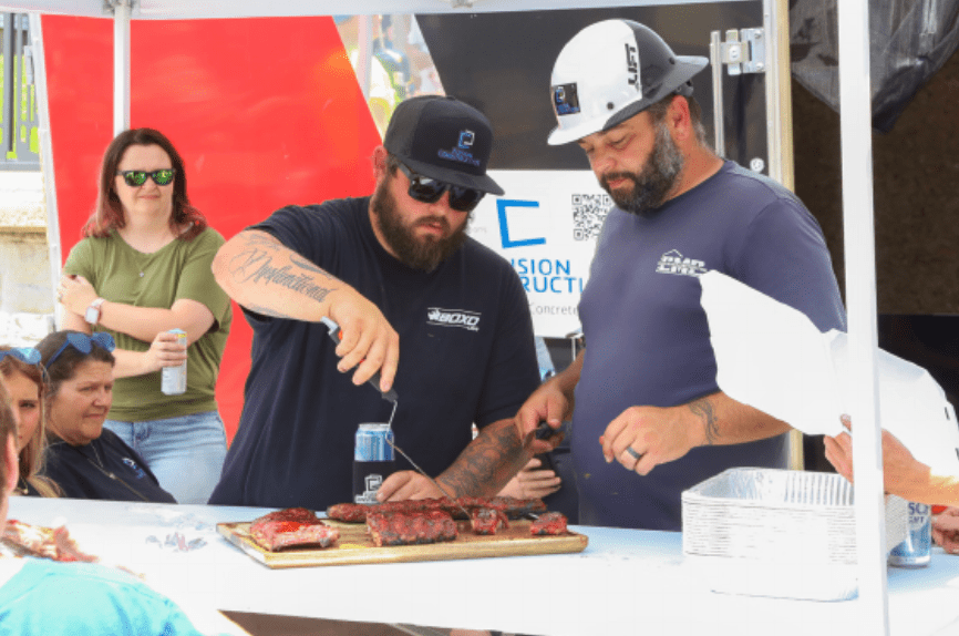Two men preparing and serving grilled meat at a food booth, with a group of people watching.