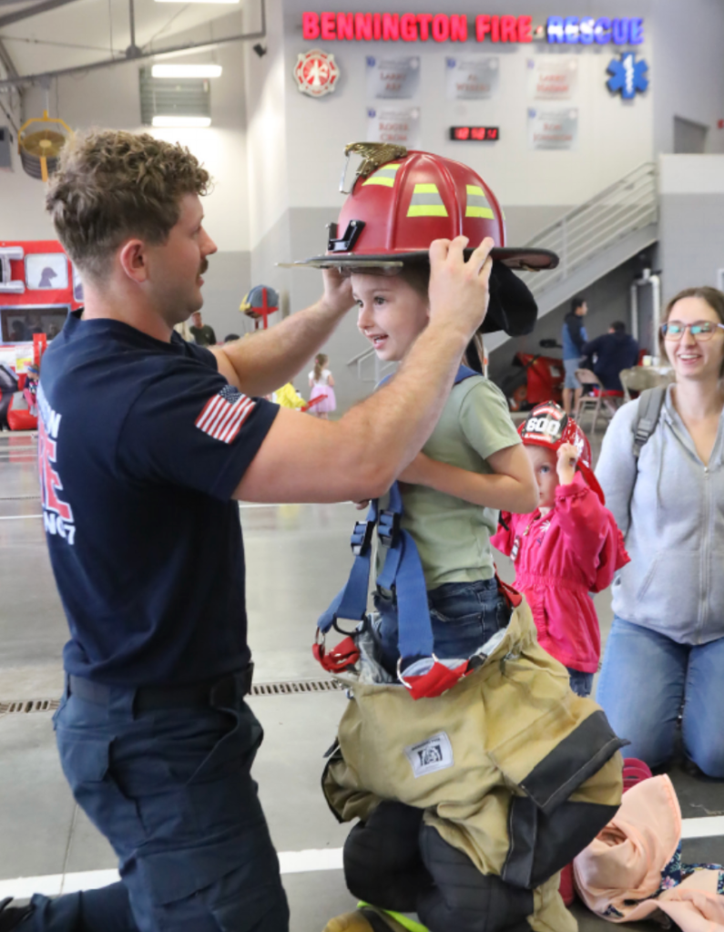 A firefighter helps a young child try on a firefighter helmet and oversized pants during a community event at a fire station.