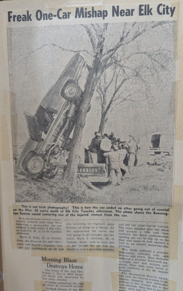 A newspaper clipping showing a car lodged in a tree after an accident near Elk City, with several individuals gathered around, assisting in the rescue of injured occupants.