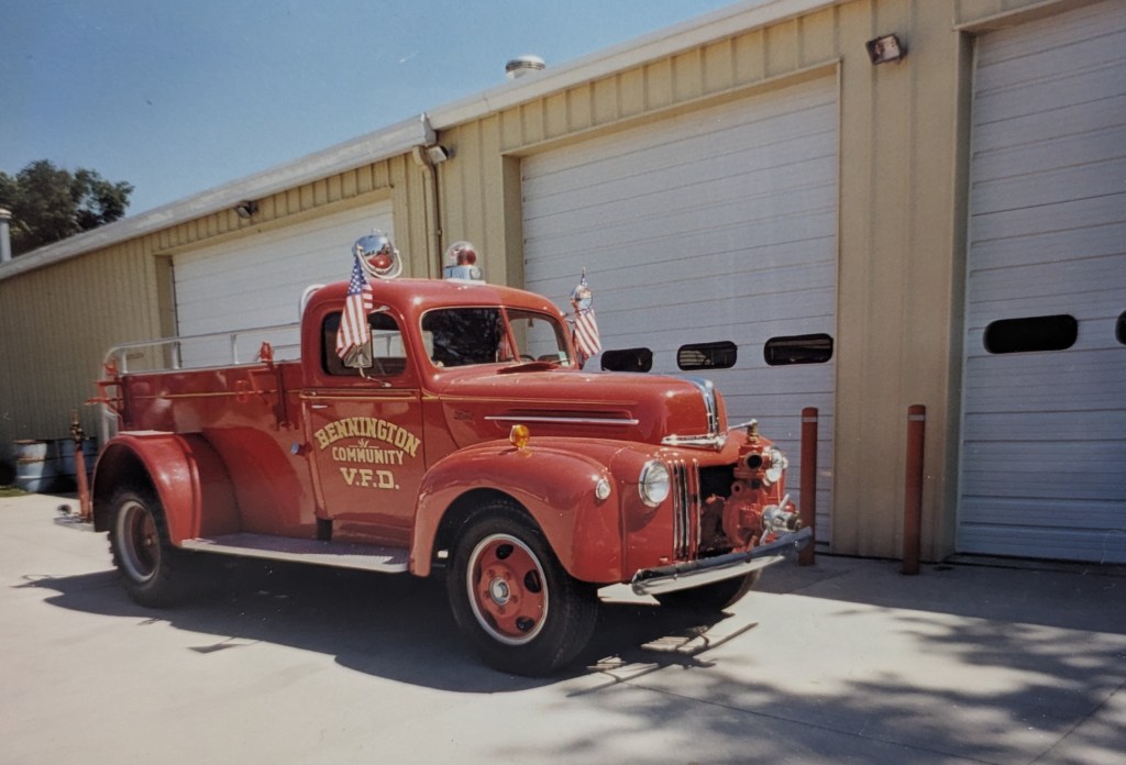 A vintage red fire truck with American flags parked in front of a fire station building.