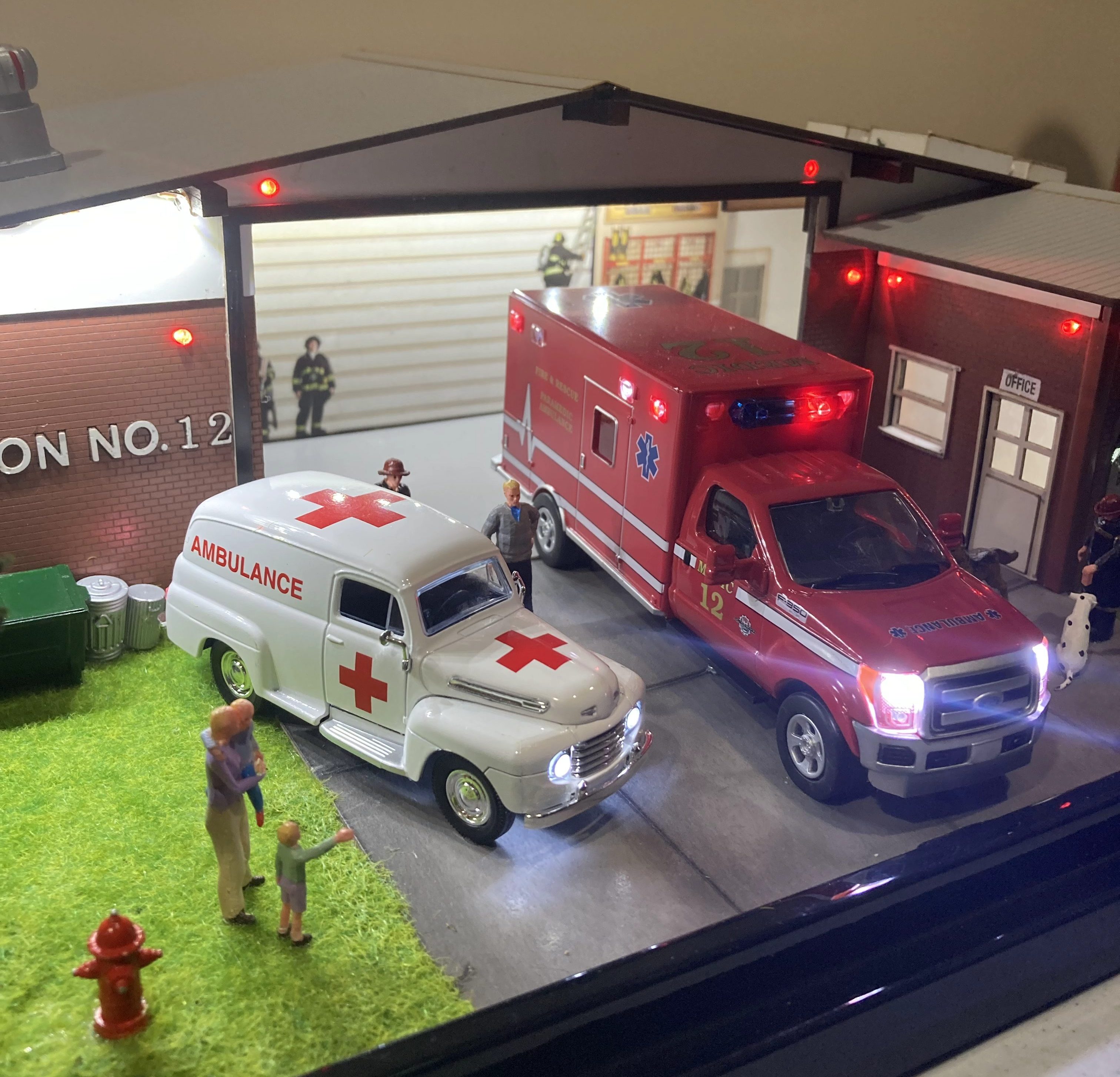 A miniature model of an ambulance and a paramedic truck parked in front of a fire station. The scene includes small figures of people and a fire hydrant, with a grassy area and details of the fire station in the background.