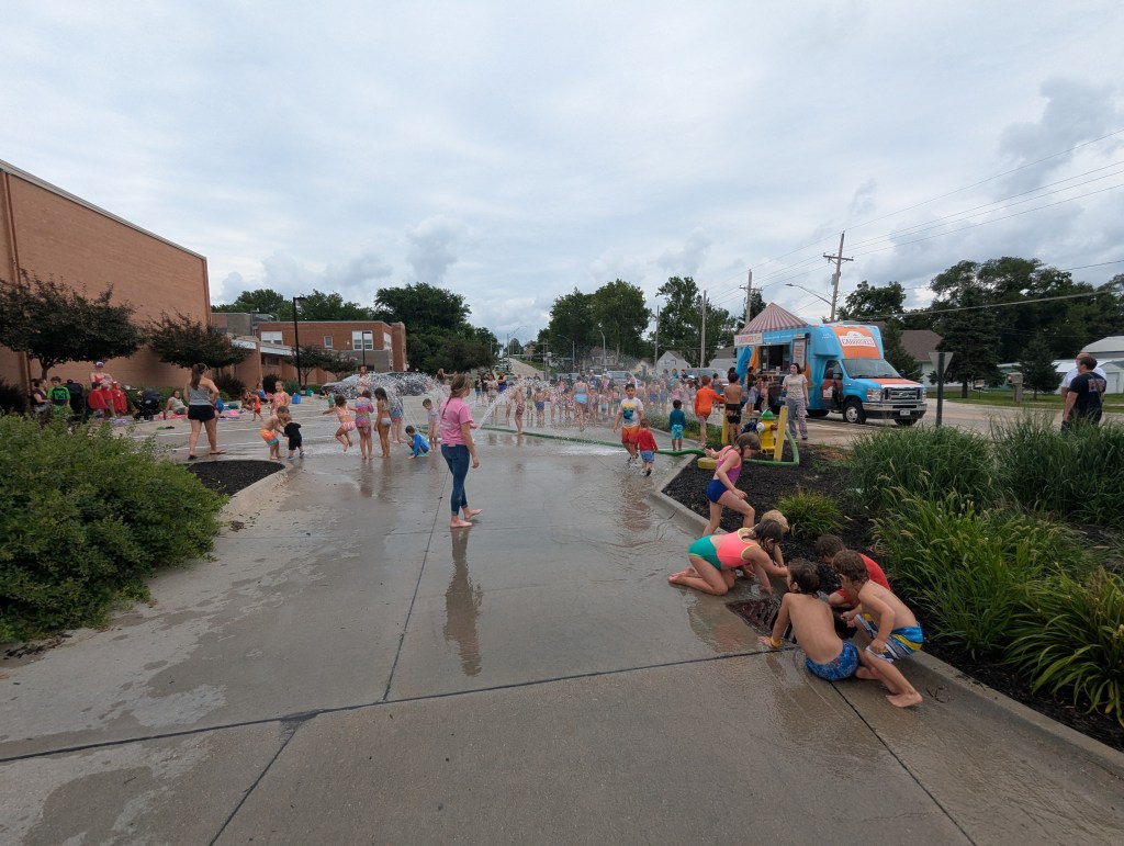 Children playing in a splash pad area with water jets, surrounded by onlookers and green foliage.