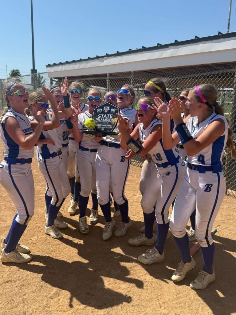 A group of young female softball players celebrating with a state championship trophy on a sunny day, wearing uniforms and sunglasses, surrounded by a dirt field.
