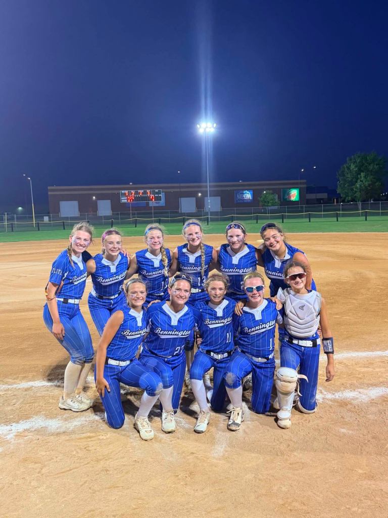 A group of young female softball players in blue uniforms posing together on a softball field at night.
