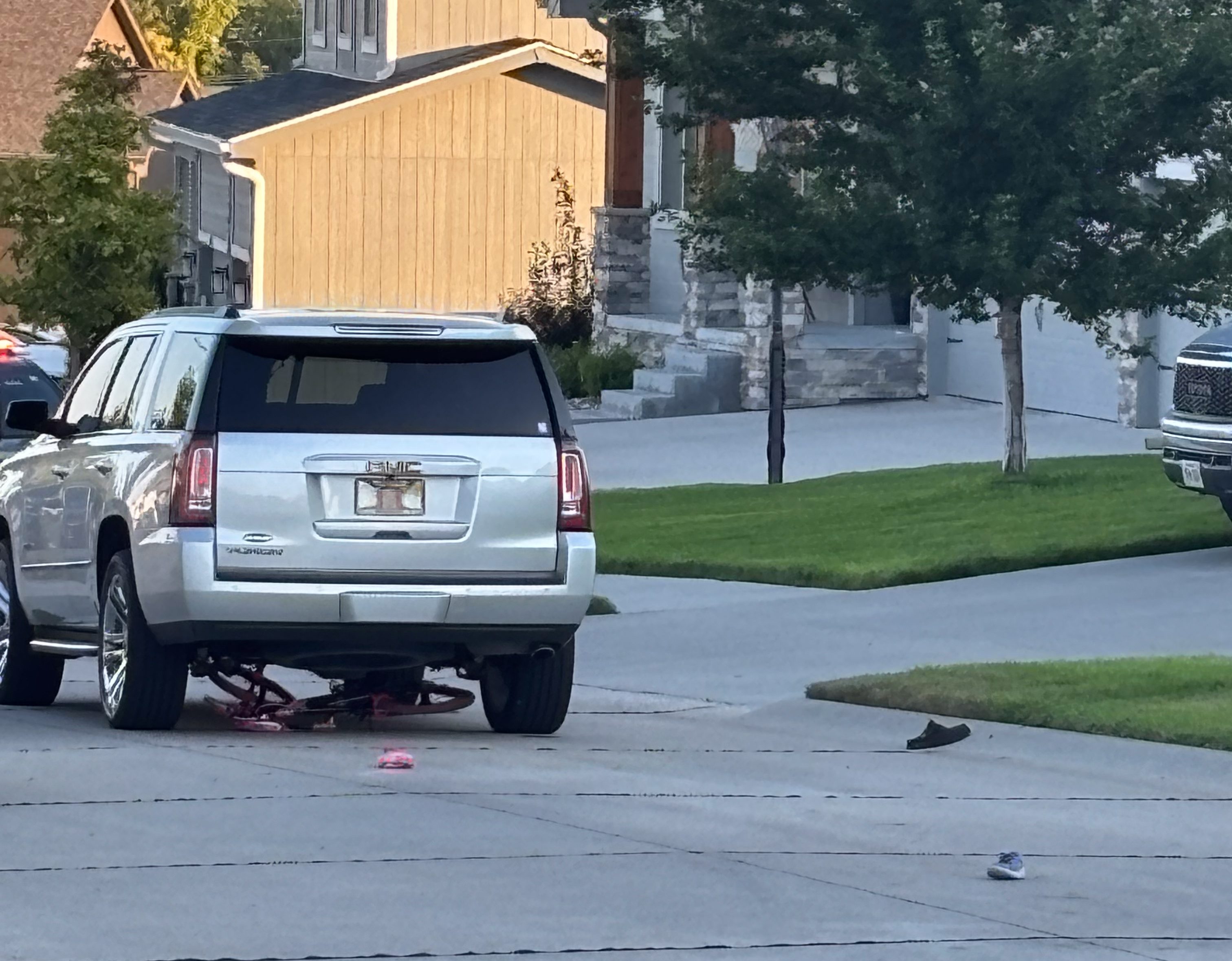 A silver SUV is lifted on a flat tire in a driveway, with a pink bicycle partially visible underneath. Two shoes are lying on the driveway. In the background, a modern house and a truck are seen.