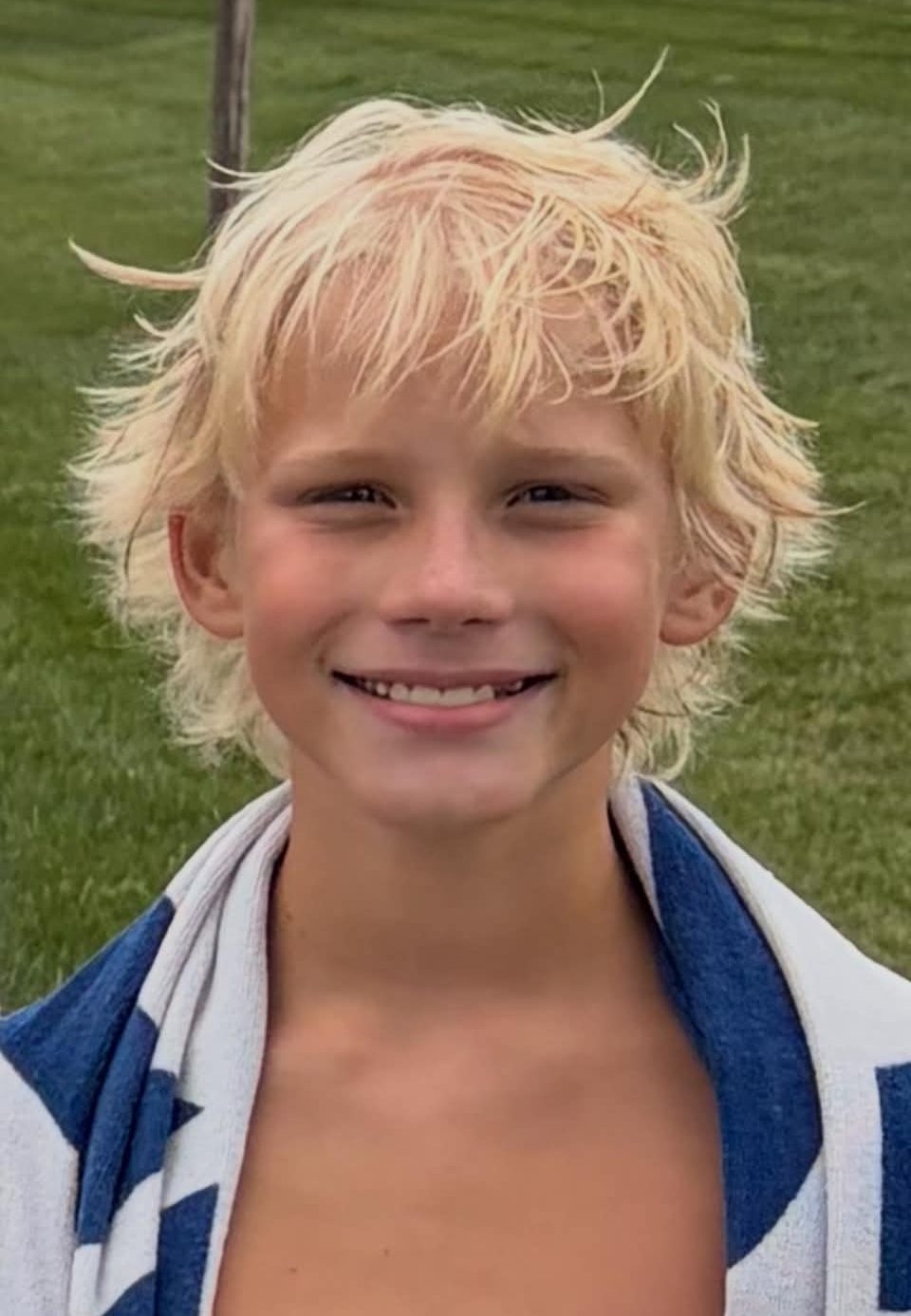 A smiling young boy with short, messy blonde hair, wearing a striped towel around his neck, standing in a grassy area.