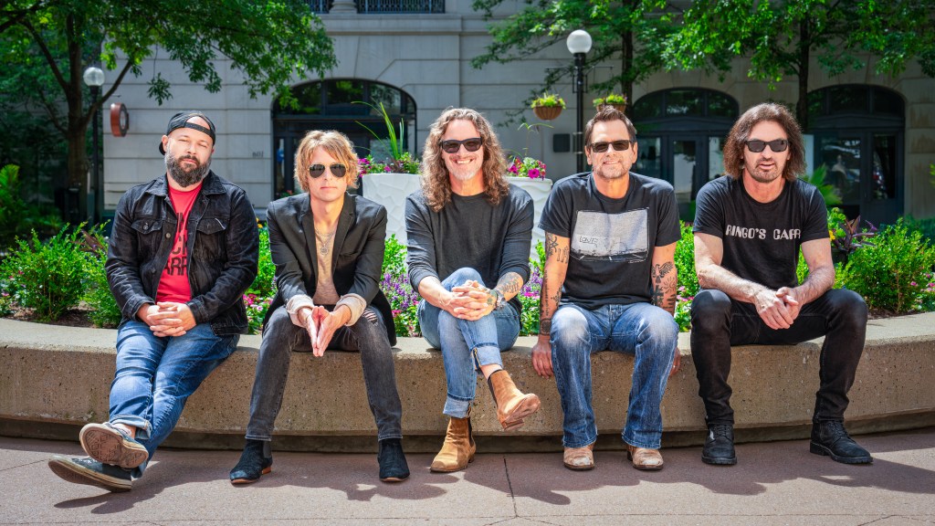 Five musicians seated on a concrete bench in a green urban setting, surrounded by flowers and trees.